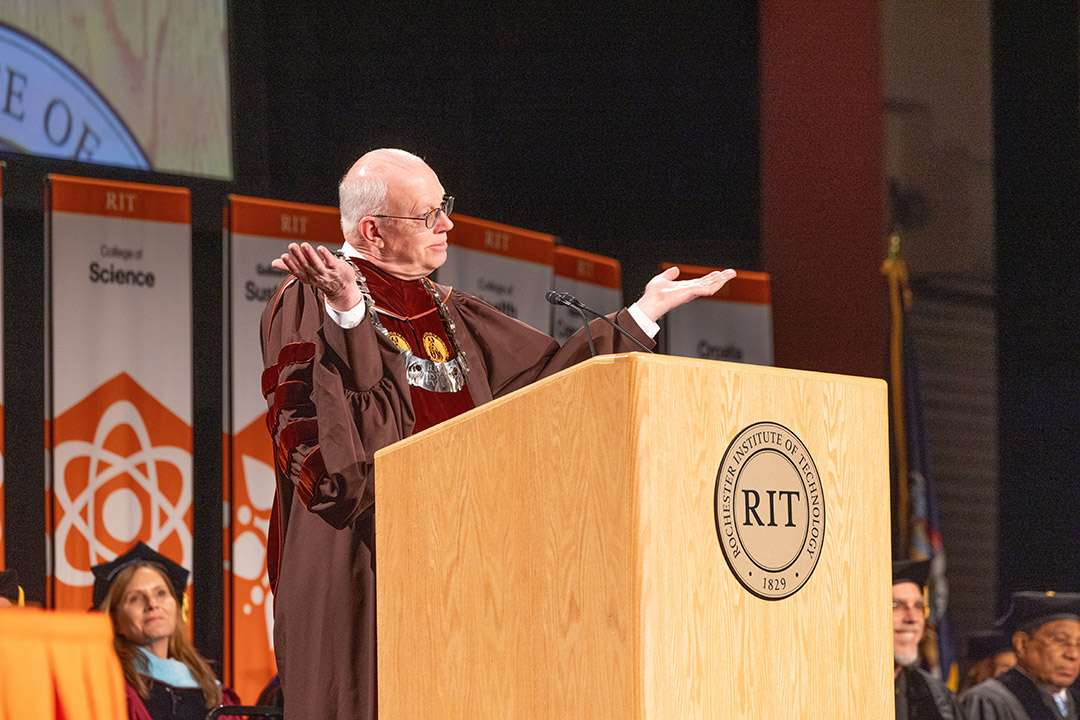 a older man in a brown acacemic robe holds his hands up in a shrug position while standing behind an R I T podium.
