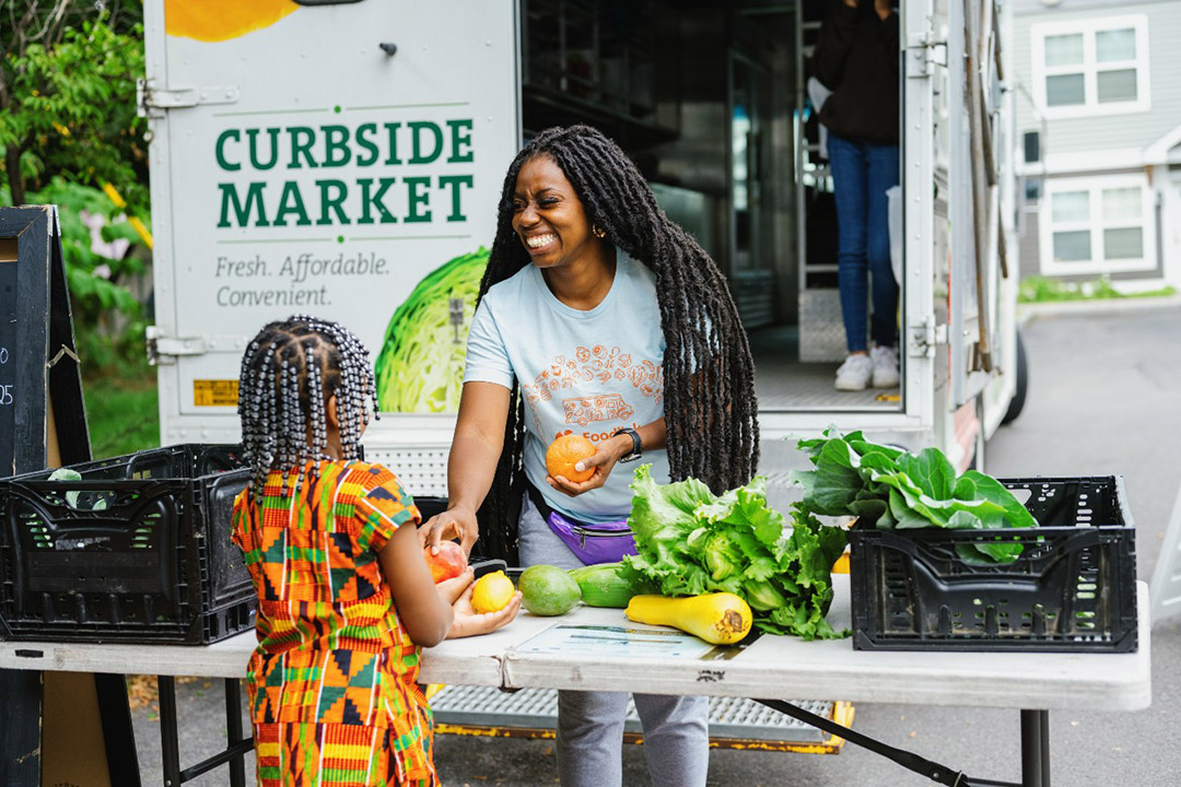 a black woman stands behind a table set up in front of the Curbside Market truck which is covered in colorful pictures of fruit and vegetables. She hands a tomato to a small black child shown only from behind.