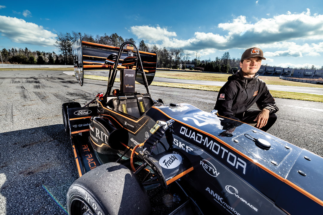a male student in a black jacket stands next to an R I T Formula S A E car on a racetrack.