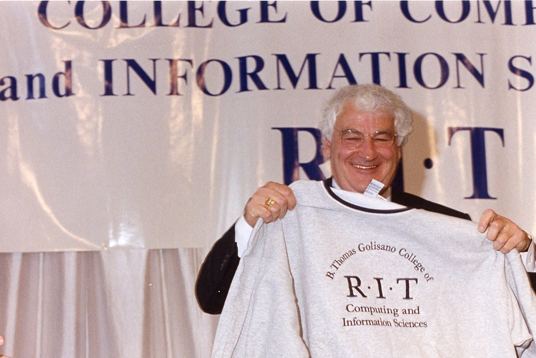 Tom Golisano stands smililing on a stage holding an R I T sweatshirt with an older style logo.