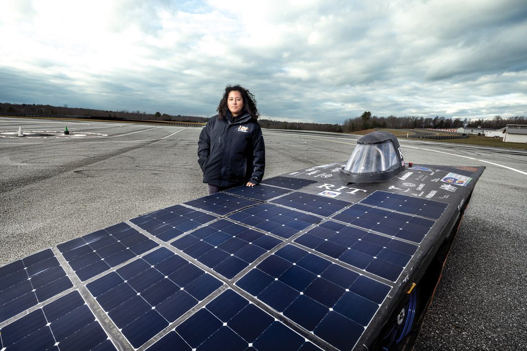 a female student stands next to a racecar built out of solar panels on a race track.