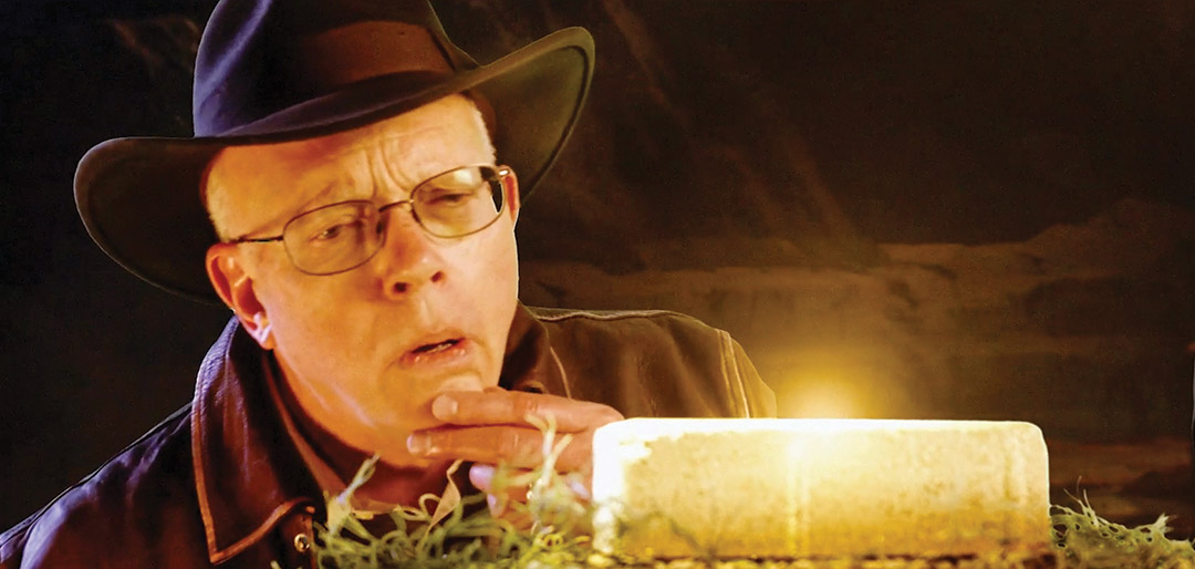 David Munson wears a leather hat and looks at a golden colored brick from the side.