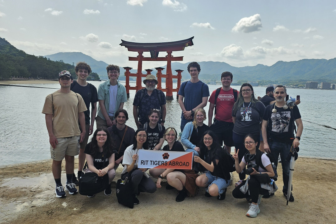 Study abroad group holding 'RIT Tigers Abroad' banner in front of the floating torii gate at Miyajima Island.