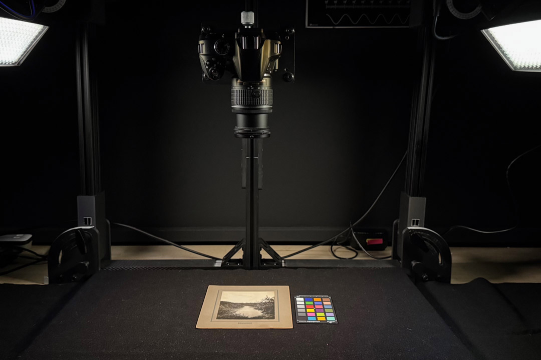 A camera mounted on a copy stand photographs a small print on a table, illuminated by two studio lights, with a color calibration card beside it.
