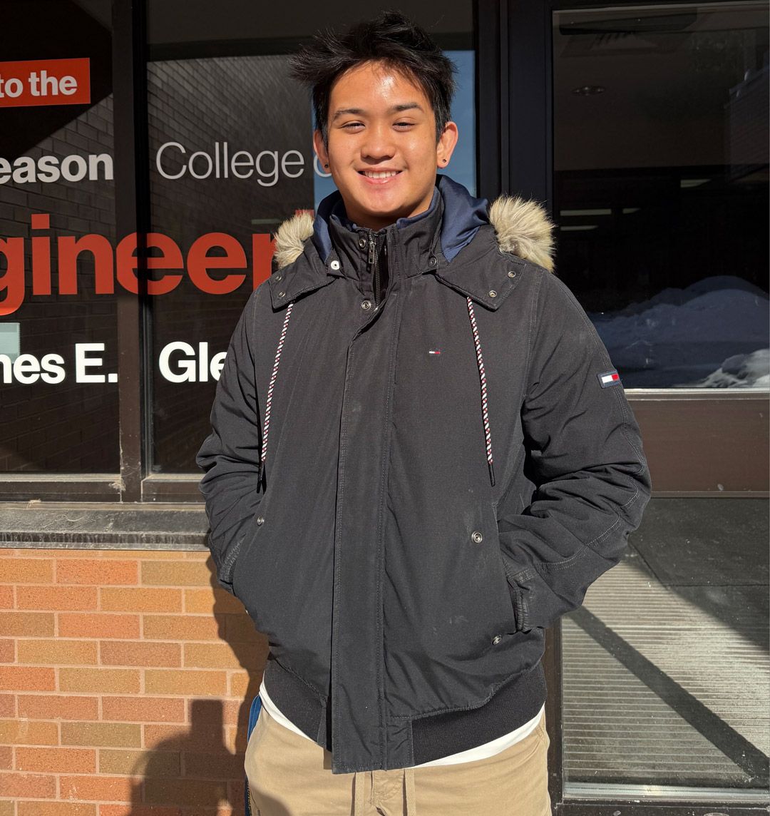 a young man in a black coat stands in front of a building with an engineering sign on the front.
