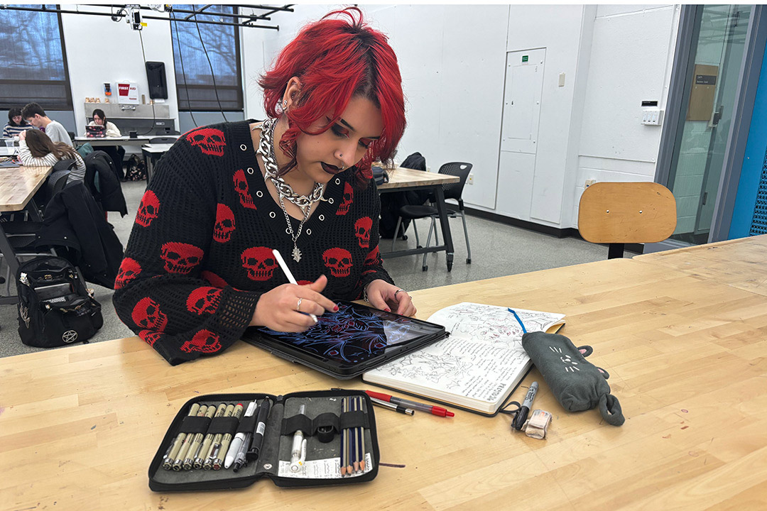A student with red hair draws on a tablet at a worktable in an art classroom, with sketchbooks and drawing tools nearby.