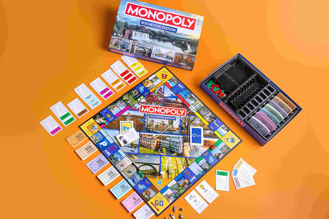 he Monopoly Rochester Edition game set up on an orange background, showing the board, property cards, Monopoly money, game pieces, and the box featuring images of Rochester landmarks.