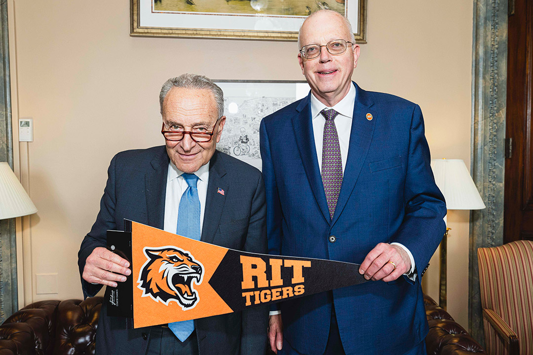 Senator Chuck Schumer and RIT President David Munson stand next to each other holding an RIT flag.