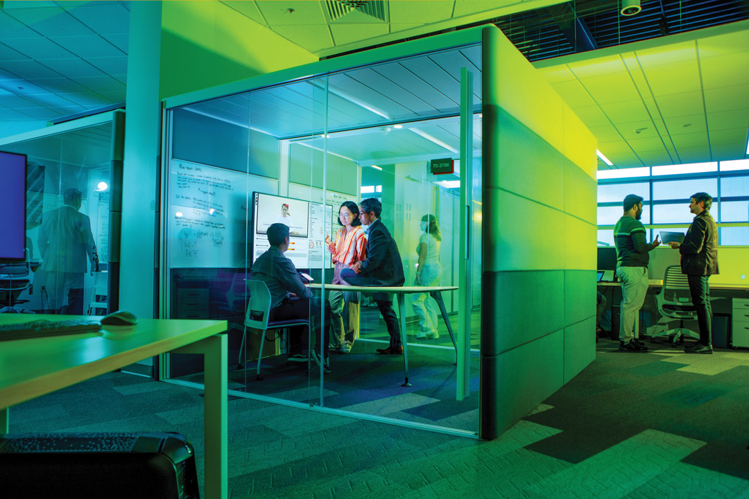 Students and a professor are seen in a glass cubicle looking at a computer screen in a modern computing lab.