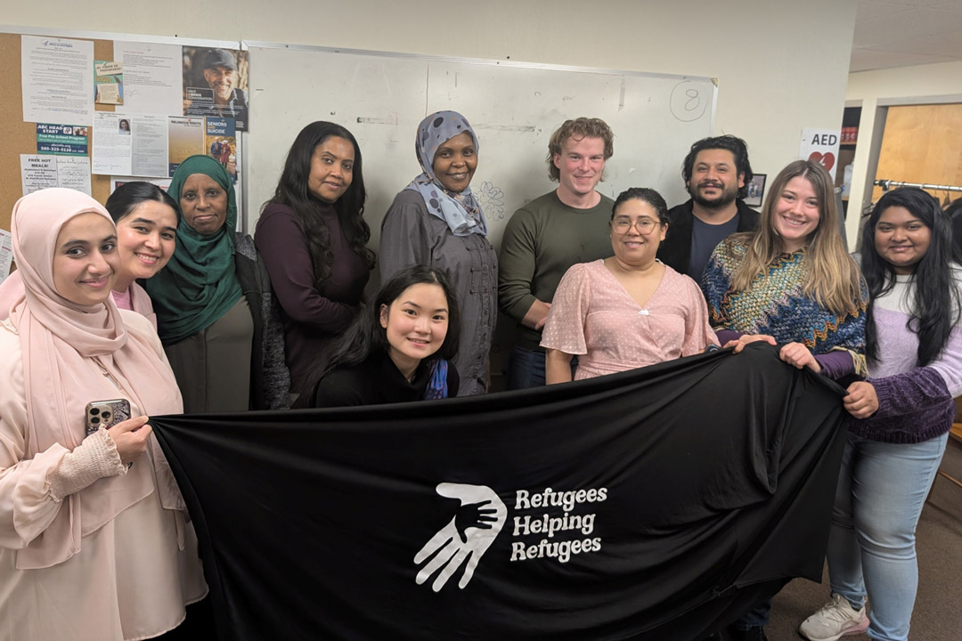 a group of students hold a black sign with white writing that reads Refugees helping refugees