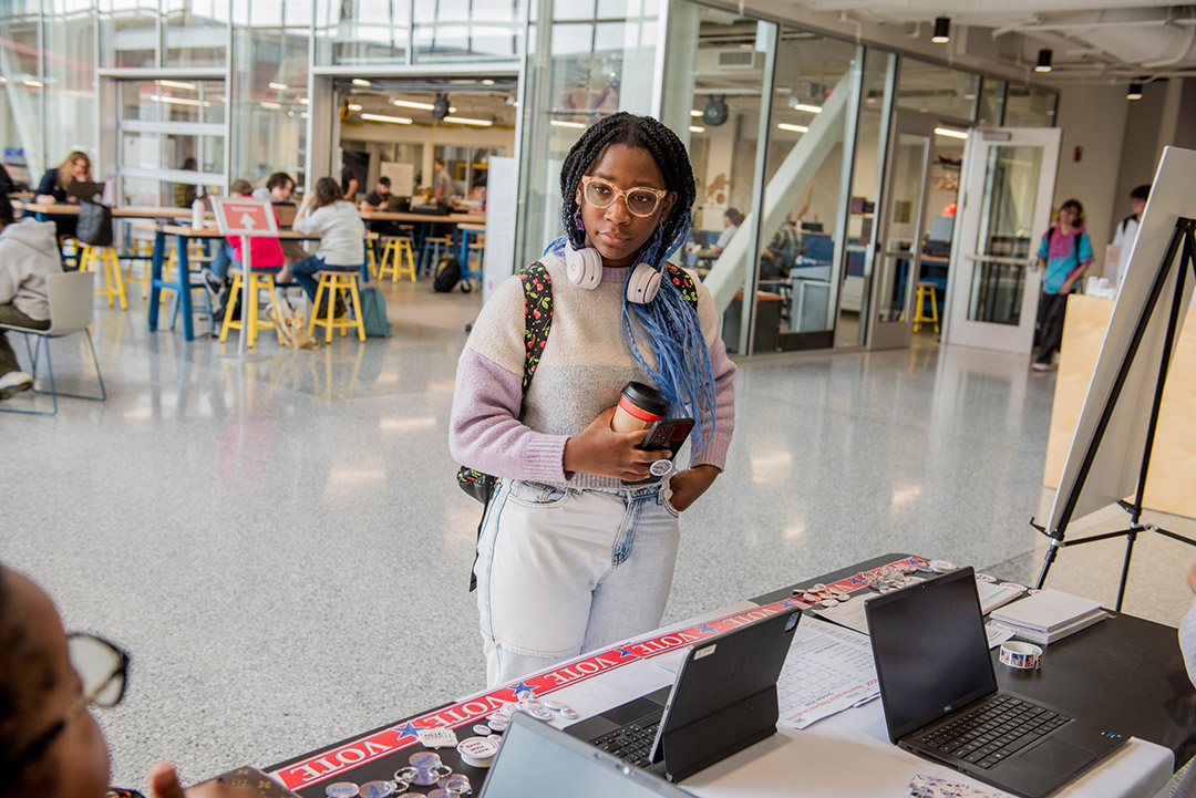 a young black woman in a gray sweatshirt and jeans stands in front of a table with information on how to vote.