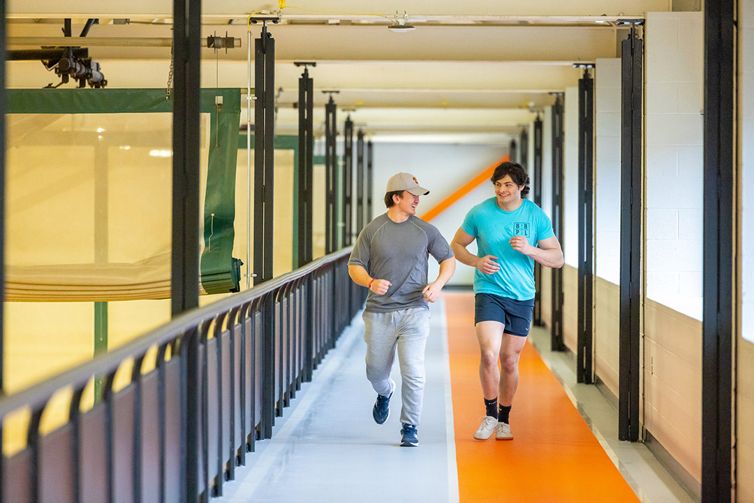 Two students jog on an indoor track at RIT, smiling and chatting as they run along the orange-and-gray lanes in a well-lit athletic facility.