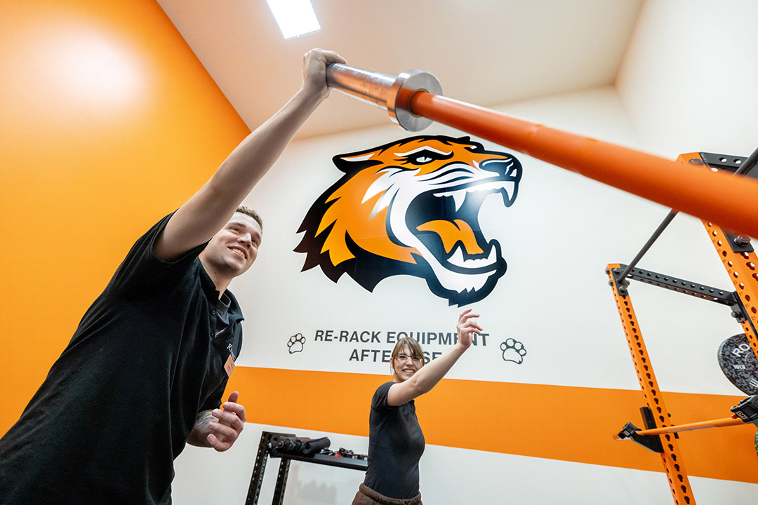 A male and female student lift an orange barbell together in a gym at RIT, smiling under a large tiger mascot mural. The phrase 'Re-rack equipment after use' is visible on the wall behind them.