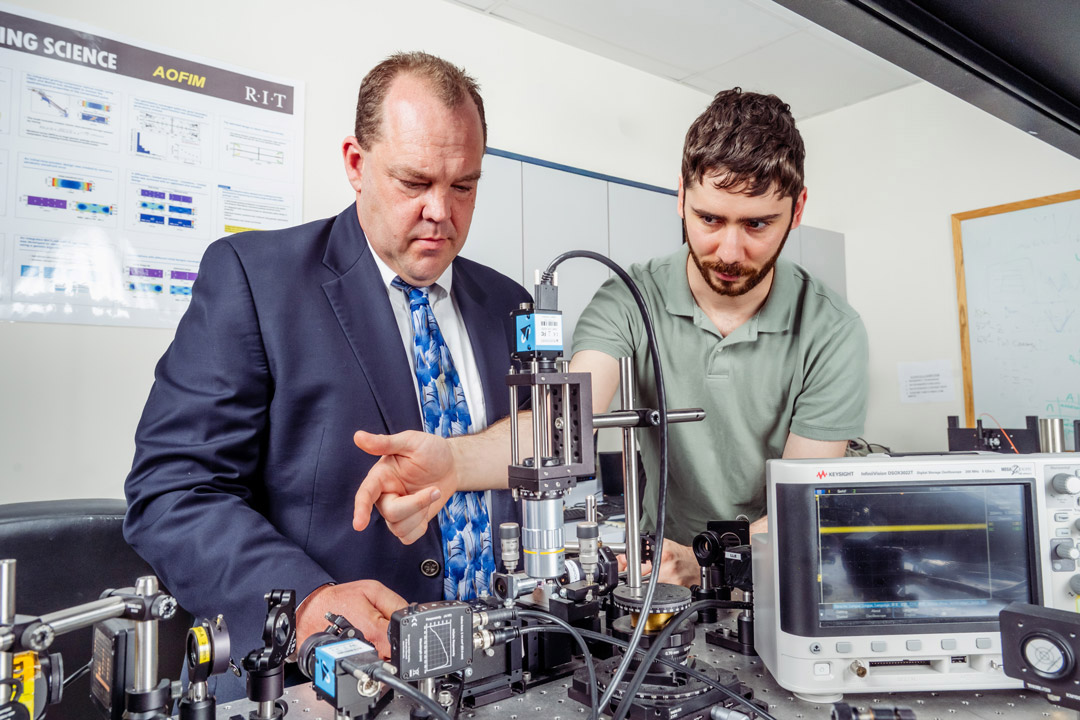 An RIT researcher demonstrates photonics laboratory equipment to a colleague.