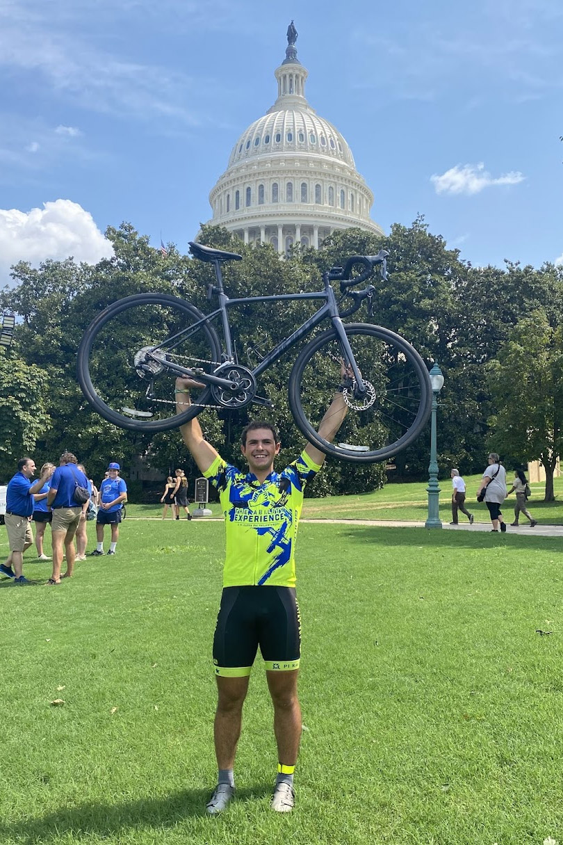 a college age male presenting person holds their bike up against the backdrop of the US capitol building.