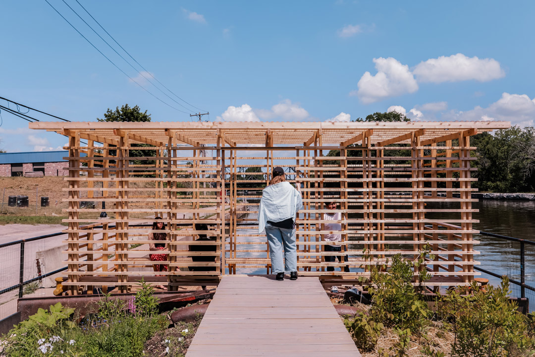 visitor walks toward a wooden pavilion built on a floating platform along the waterfront.