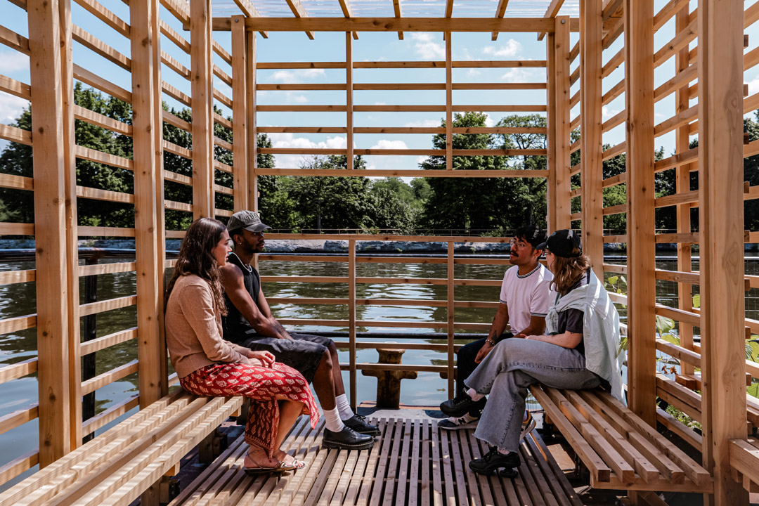 Four people sit inside a wooden floating pavilion overlooking the water.