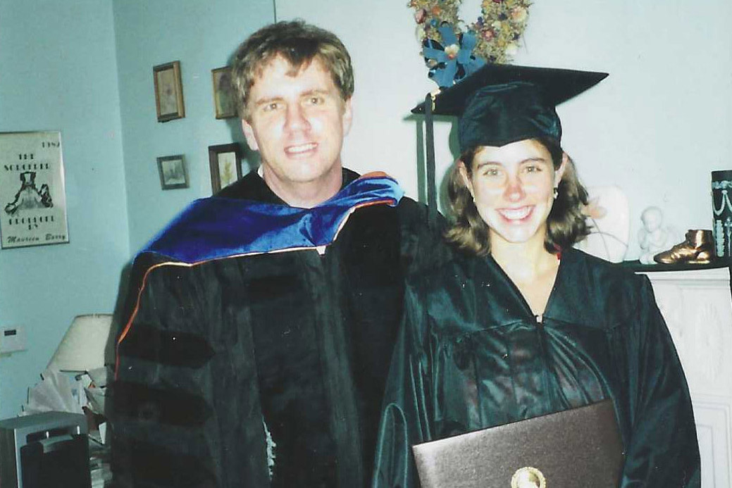 a man and a young woman stand in a livingroom. The woman holds a diploma from college.