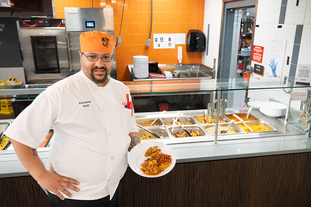 a man in an orange chef hat stands next to a glass barrier with food behind it. ALTTEXT