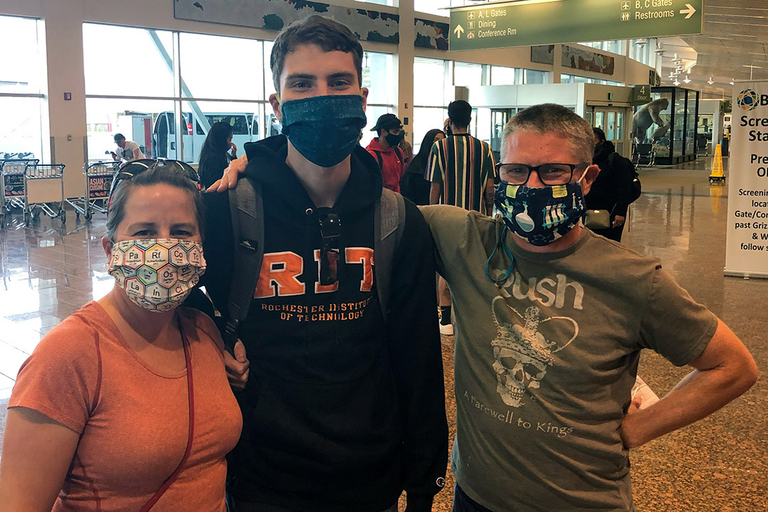 a student in an RIT sweatshirt stands between two adults in the Rochester airport arrivals area.