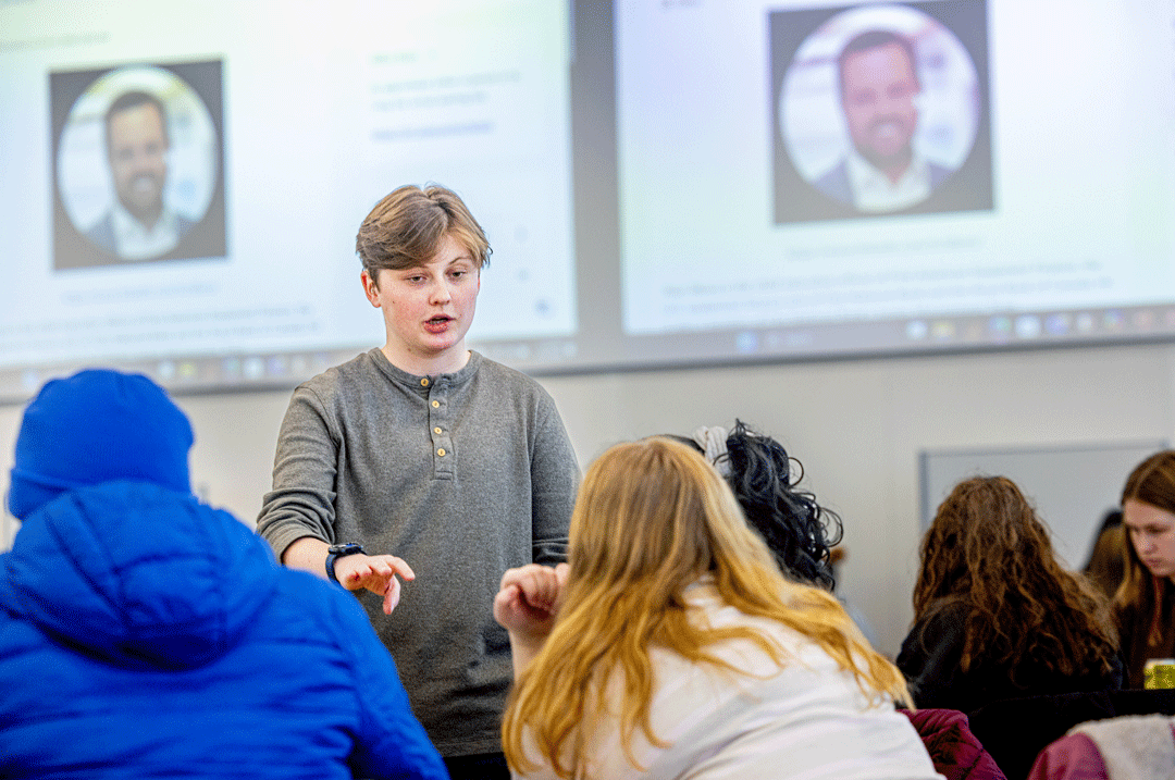 Standing student teacher speaks to a group of classmates in a classroom 