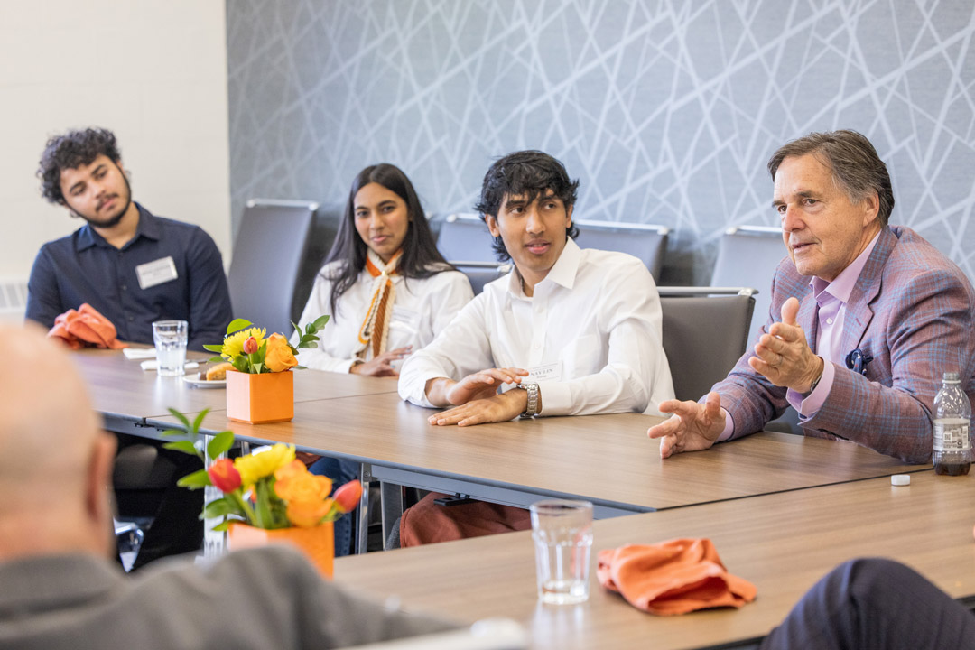 Three students sit at a table listening to a man in a suit talking.