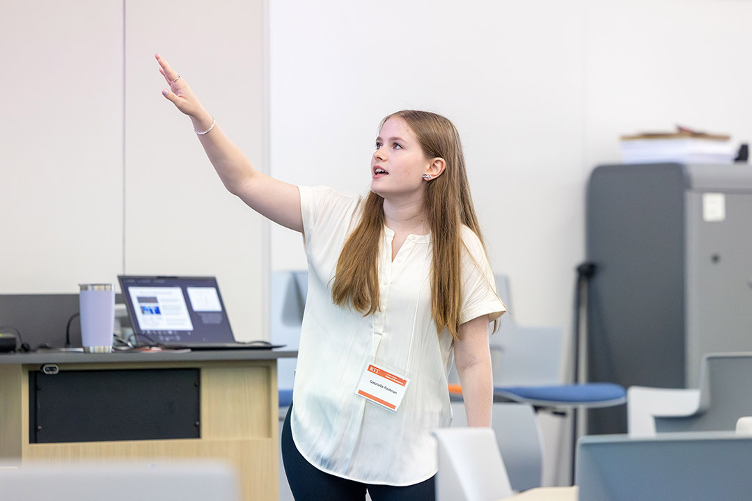 Young woman in a white blouse raises her hand while presenting beside a laptop.