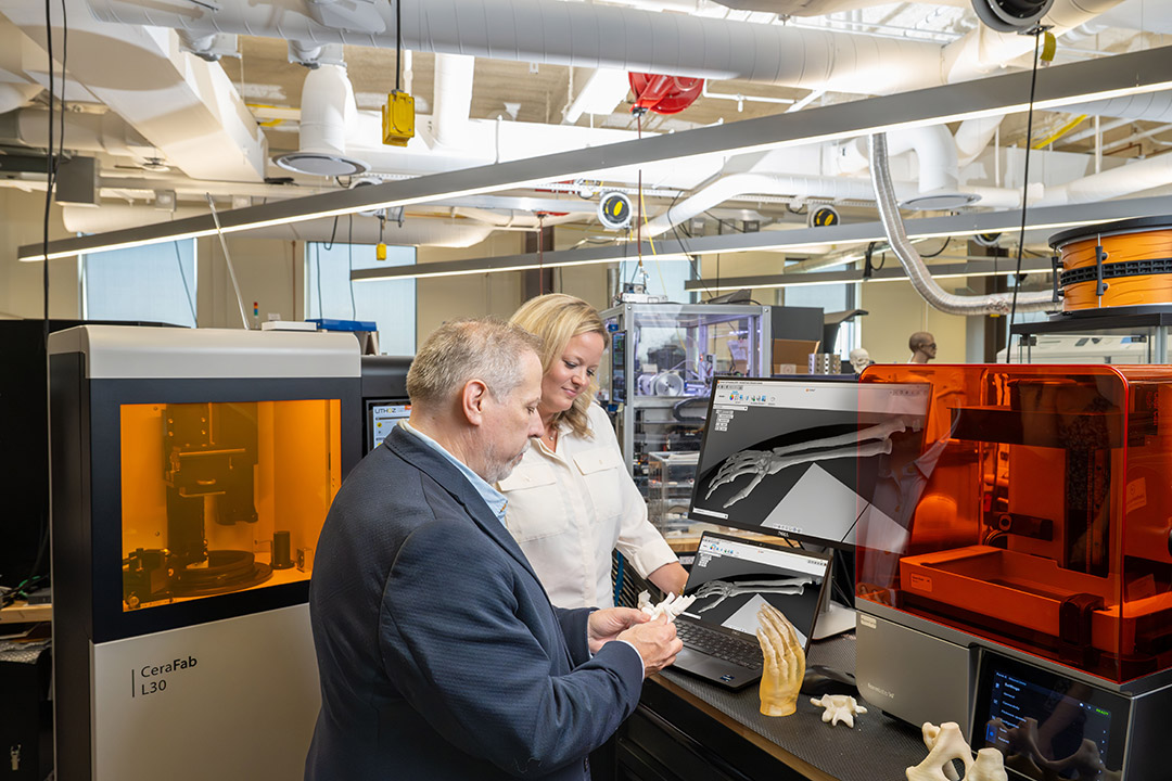 ngineer and colleague examine a 3D-printed bone model alongside its CAD view on a monitor.