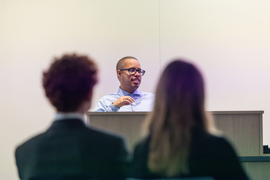 Male speaker with glasses addresses two blurred audience members from behind a lectern.