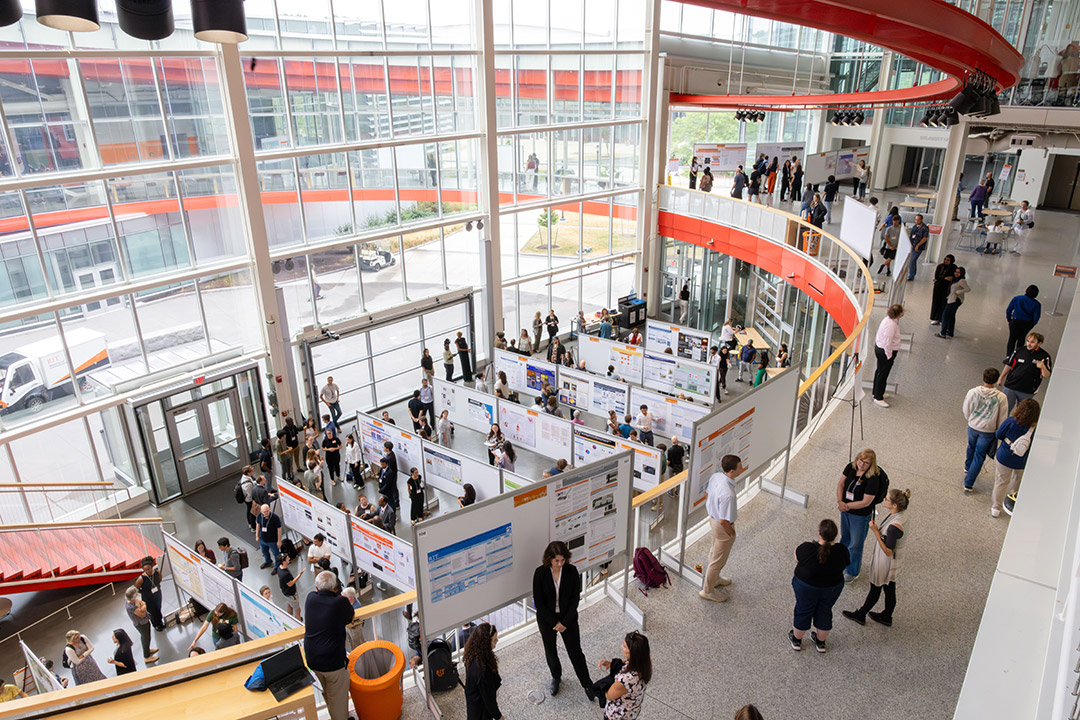 Attendees walk and talk beside posters along a curved glass-walled atrium.