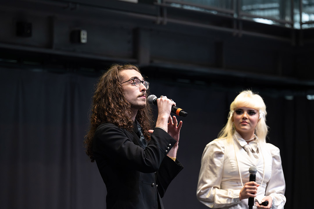 Man and woman holding microphones speak on stage with dark curtain backdrop.