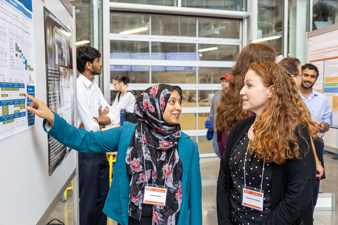 Woman in a floral hijab points at a poster chart as a colleague listens.