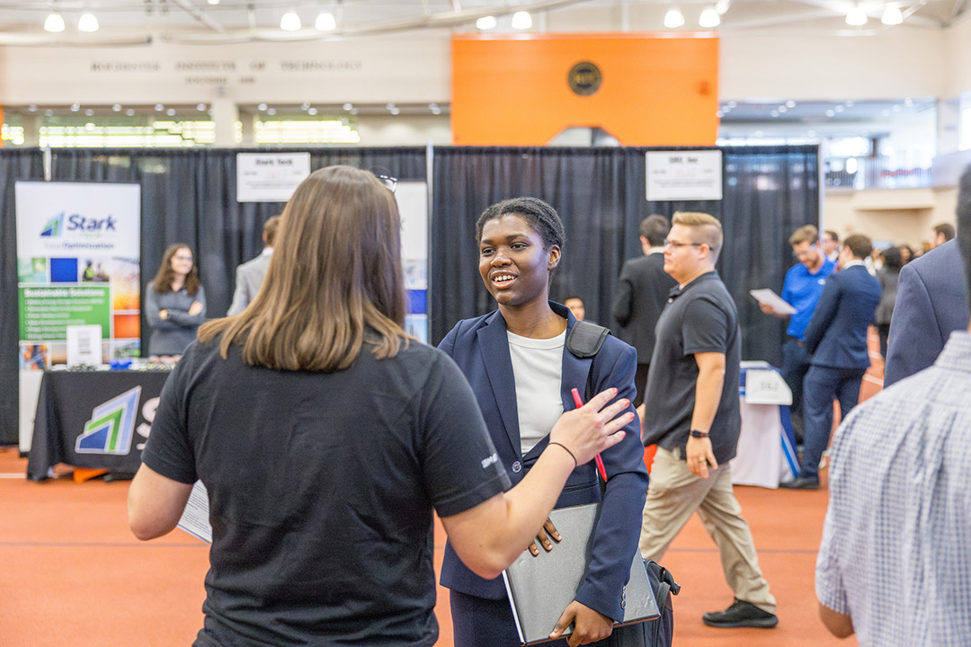a female student faces the camera speaking to a woman in a black tshirt, seen only by her back.