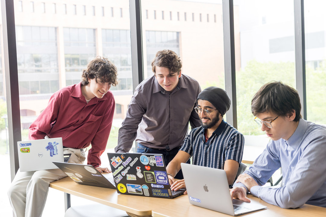 Four college age males sit at a table working together with laptops in a sunny room on a campus