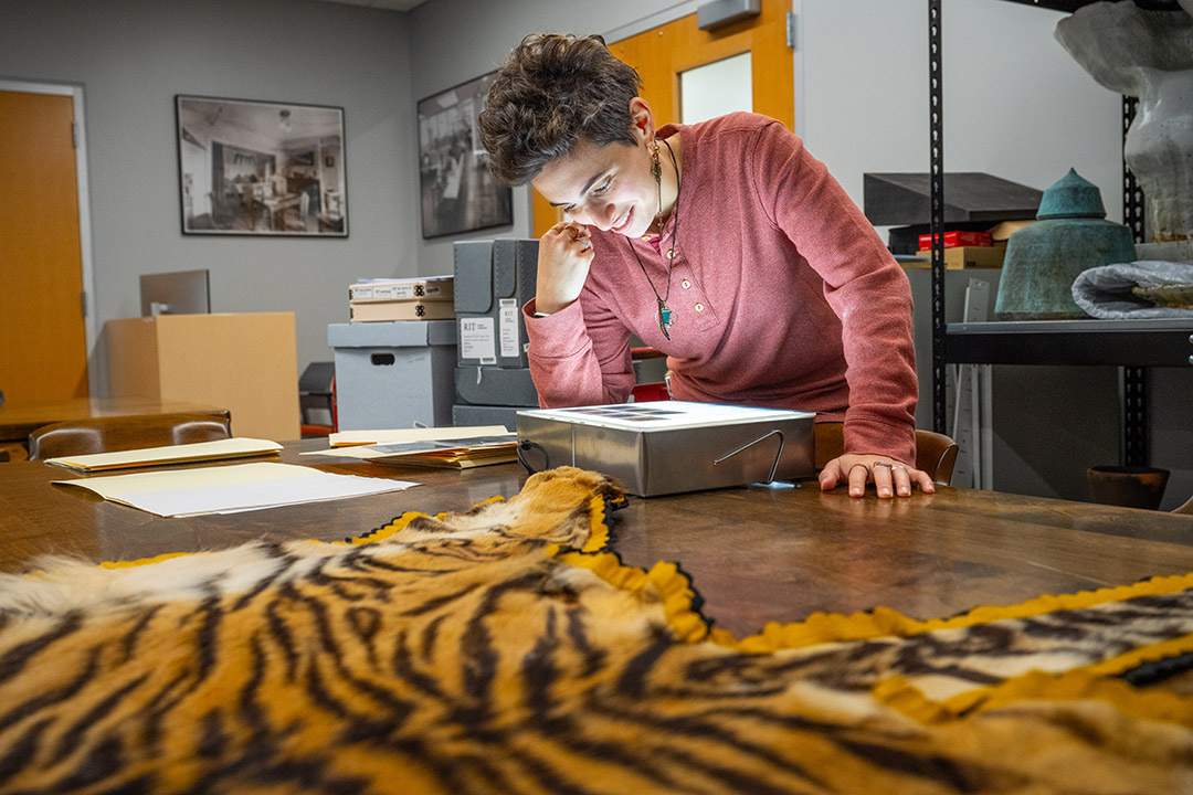 a woman with short dark hair leans over a lightbox on a table looking at slides in a research repository. In the foreground, the pelt of a tiger is resting on the table.