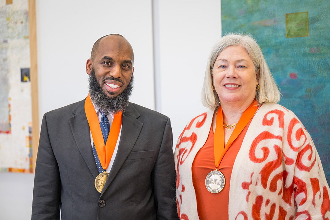 a young black man, Irshad Altheimer, and and an older white woman, Ellen Granberg, stand next to each other smiling. The man wears a black suit and a bronze medal hangs arround his neck.