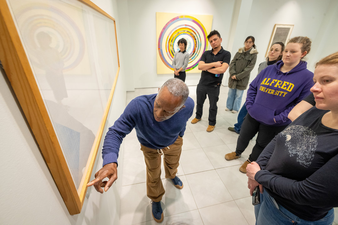a man bends down pointing at the bottom of a piece of art as students look on.