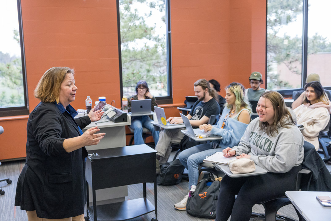 a woman stands in front of a class speaking to them while they interact