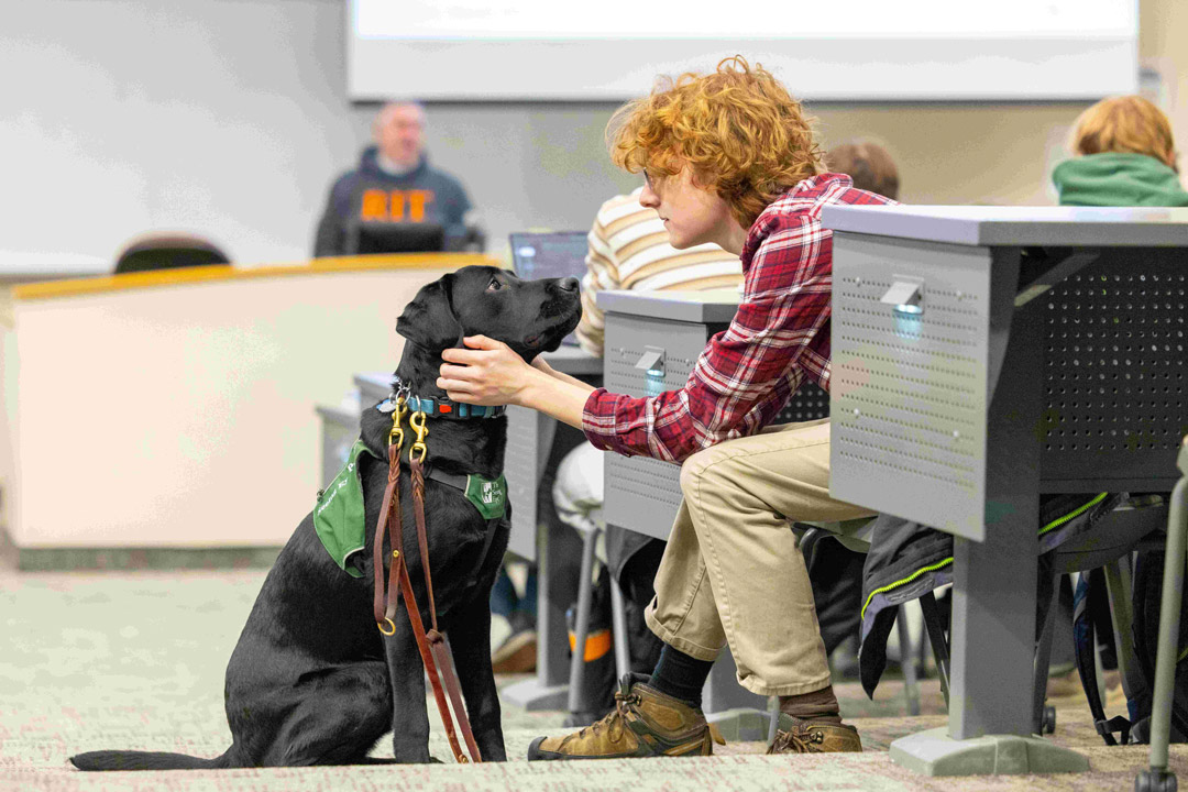 a man with red hair sits in a lecture hall with a black dog, holding the dogs face between his hands in a loving manner.