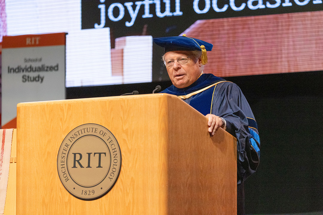a man stands a an R I T podium addressing an audience