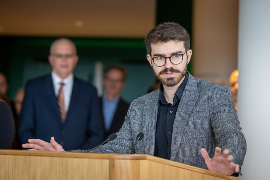 a young man in a gray suit stands at a podium