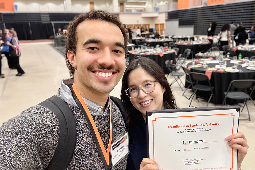 TJ Hampton, left, and associate professor Hye-Jin Nae, right, pose with Hampton's Excellence in Student Life Award. Hampton is holding the camera in selfie-mode and Nae is holding Hampton's award. Both of them are smiling.