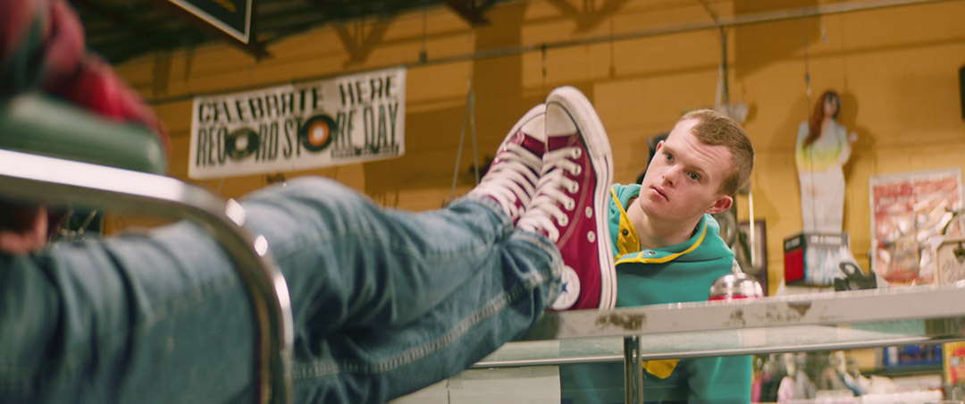 a mans legs, clad in denim jeans, are lifted up on to a table with red converse. A man sits behind the same table.