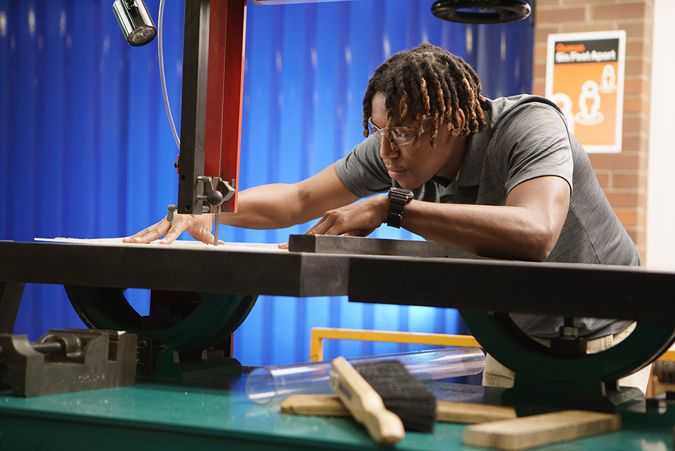 An RIT student, wearing safety glasses and a gray collared shirt, working on a band saw in the Machine Shop.