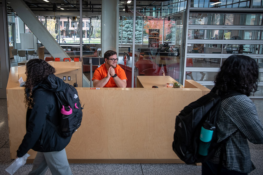 college student sitting behind an information desk as other students walk by.