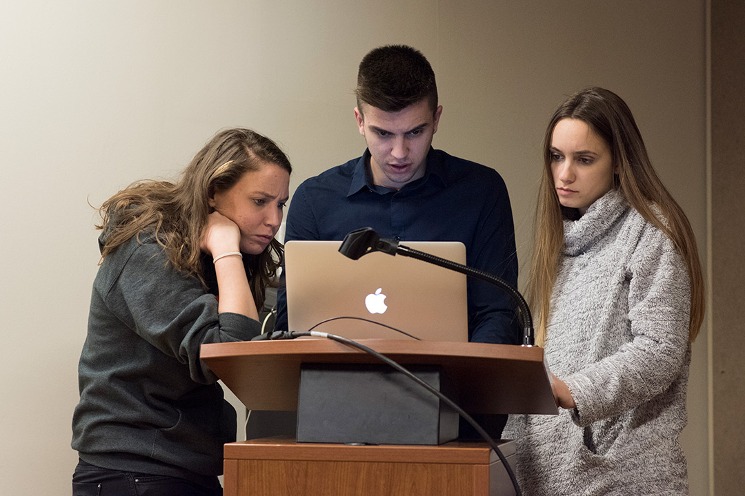 Three student standing at podium looking at laptop