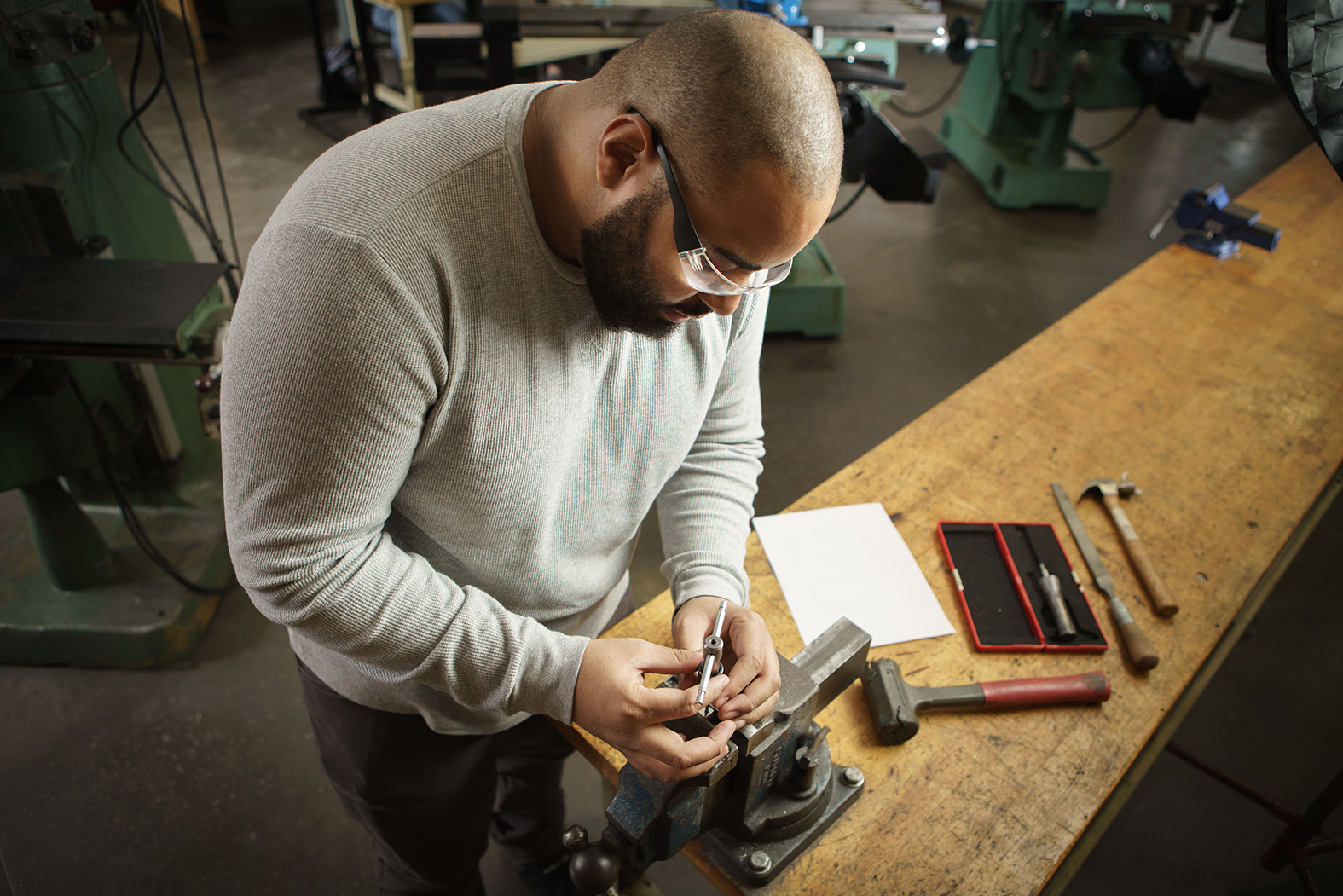 A student engineer, wearing safety glasses and a gray shirt, working with a clamp in the Machine Shop.