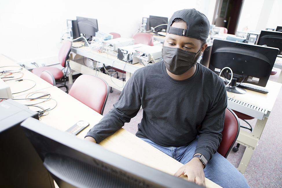 A college student wearing a backwards baseball cap and black shirt, working on a desktop computer in an electrical engineering lab.