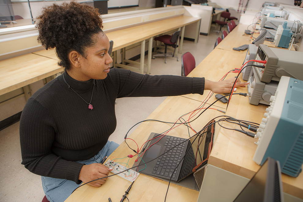 An RIT student, Roxette Burgos wearing a black turtleneck sweater, working in an electrical engineering lab.