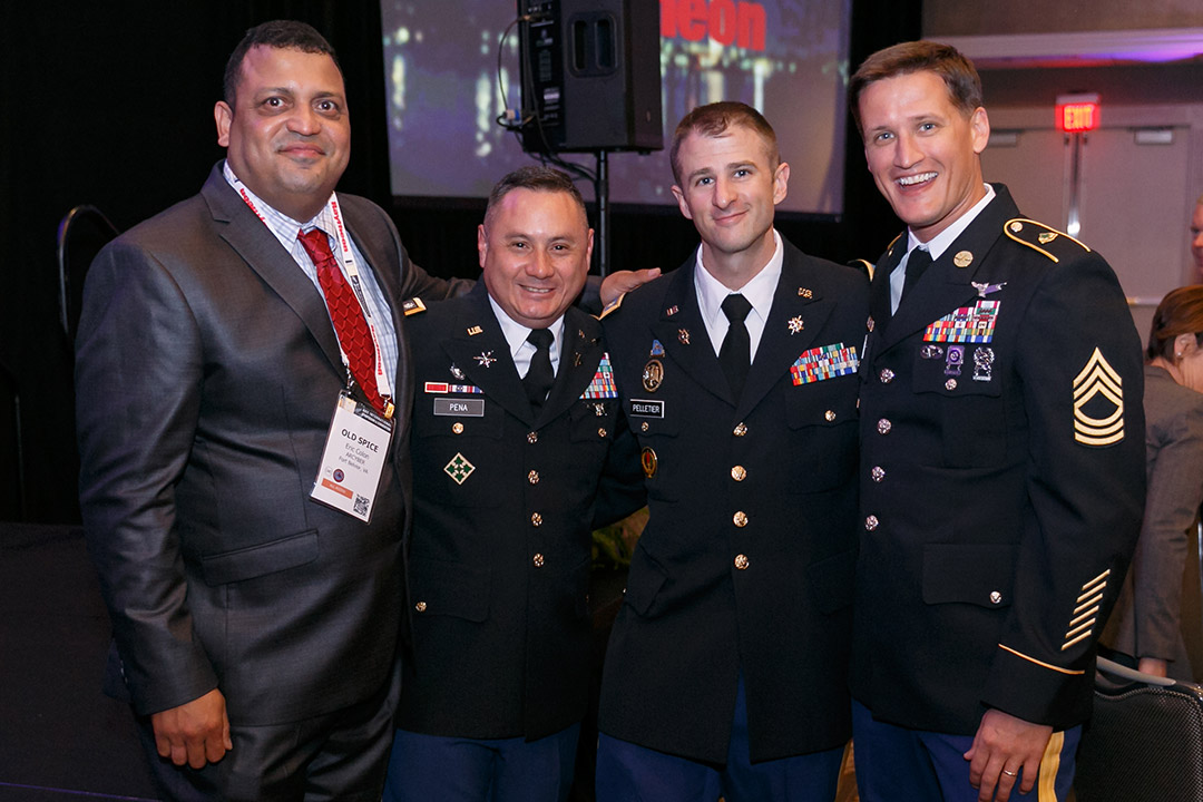 four people posing for a photo, one wearing a suit and the others wear military dress uniforms.